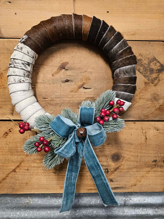 Cowhide wreath with natural elements, a blue ribbon, and a bell against a rustic wood plank background.