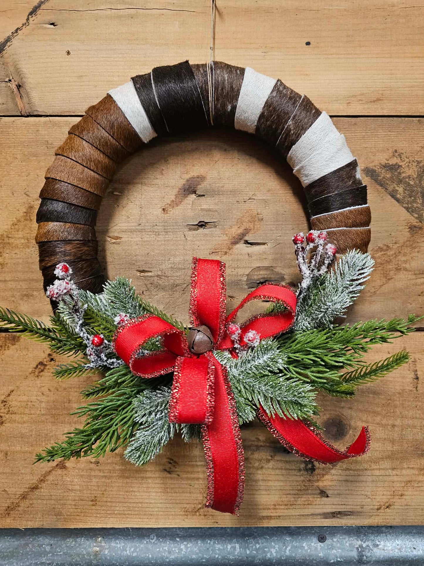 Cowhide wreath with greenery, a red ribbon, and a bell against a rustic wood plank background.