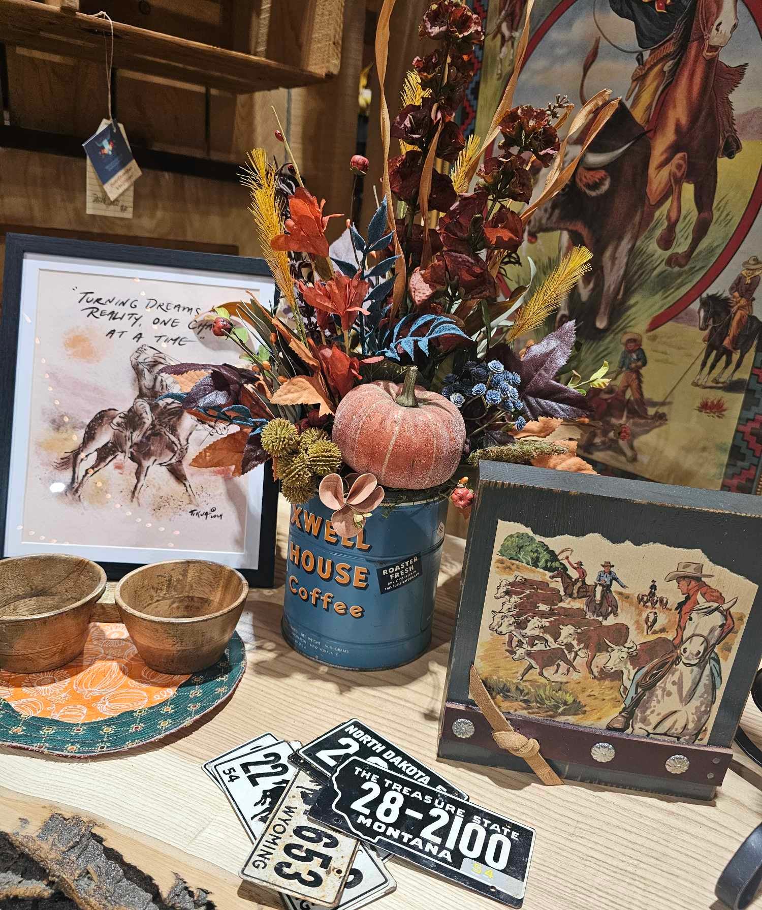 Artwork on a table, including a fall flower arrangement with a small pumpkin, a cowboy decoupage, and antique license plates.
