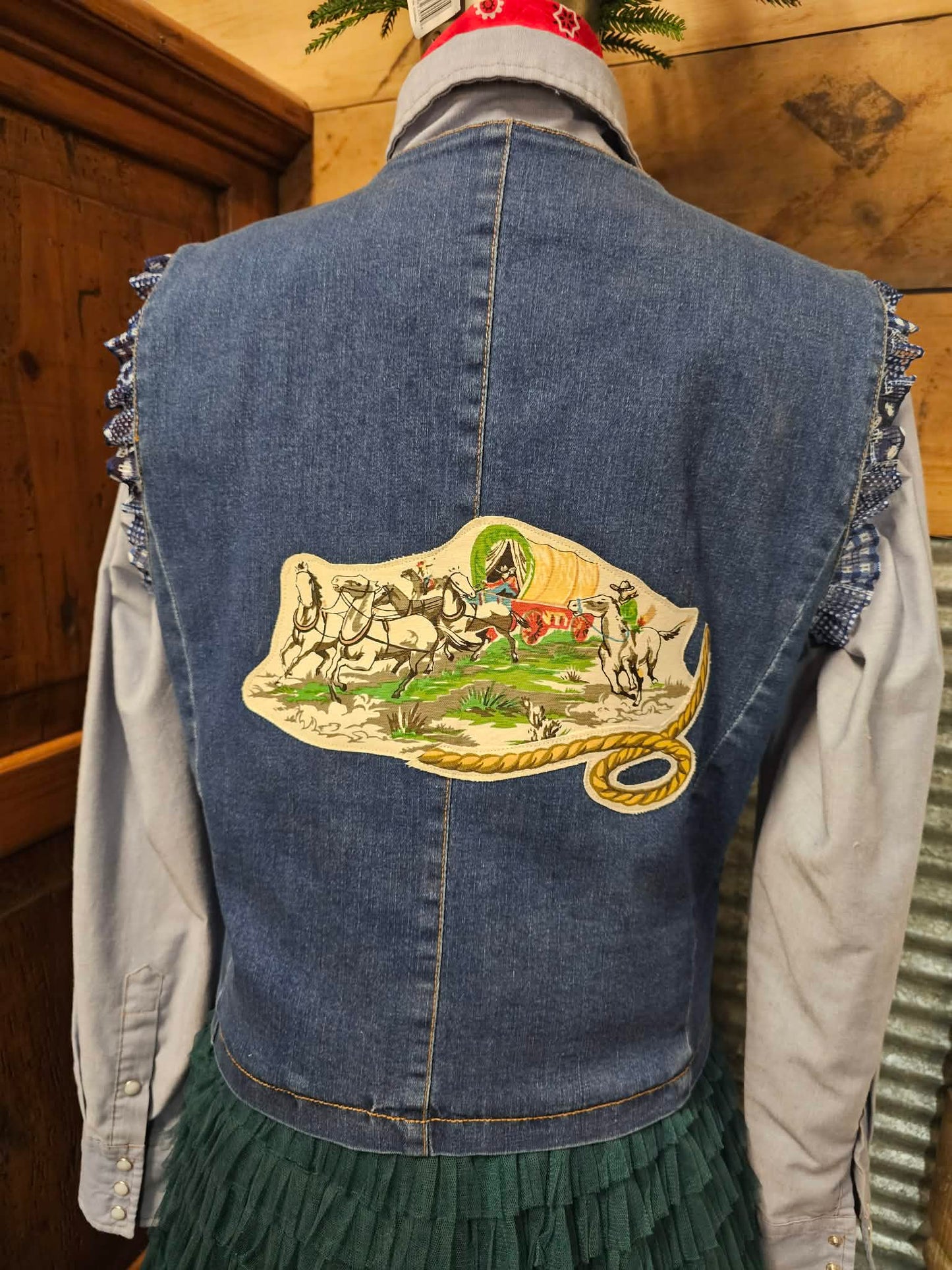 Denim vest with a colorful appliqued design on the back, displayed against a wooden wall.