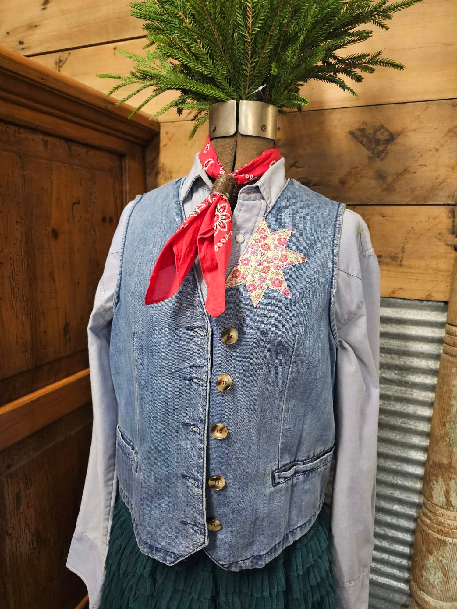 Mannequin wearing a denim vest with a floral shirt and red bandana, against a wooden background. Vest has a floral star quilted on the left lapel. 