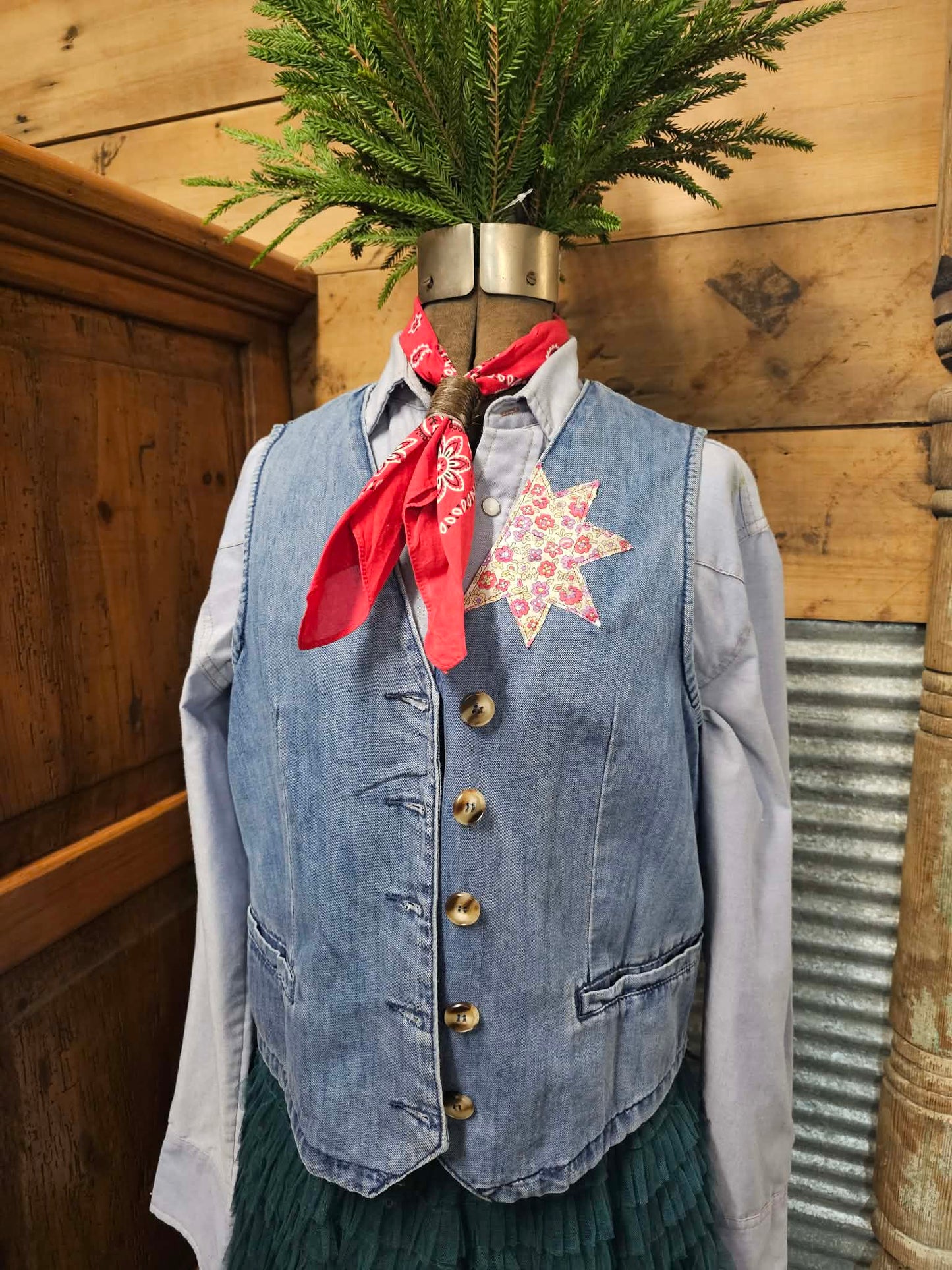Mannequin wearing a denim vest with a floral shirt and red bandana, against a wooden background. Vest has a floral star quilted on the left lapel. 