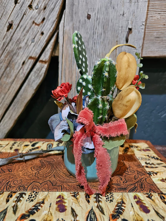 Decorative arrangement with cactus, flowers, and a pink bow on a patterned surface.