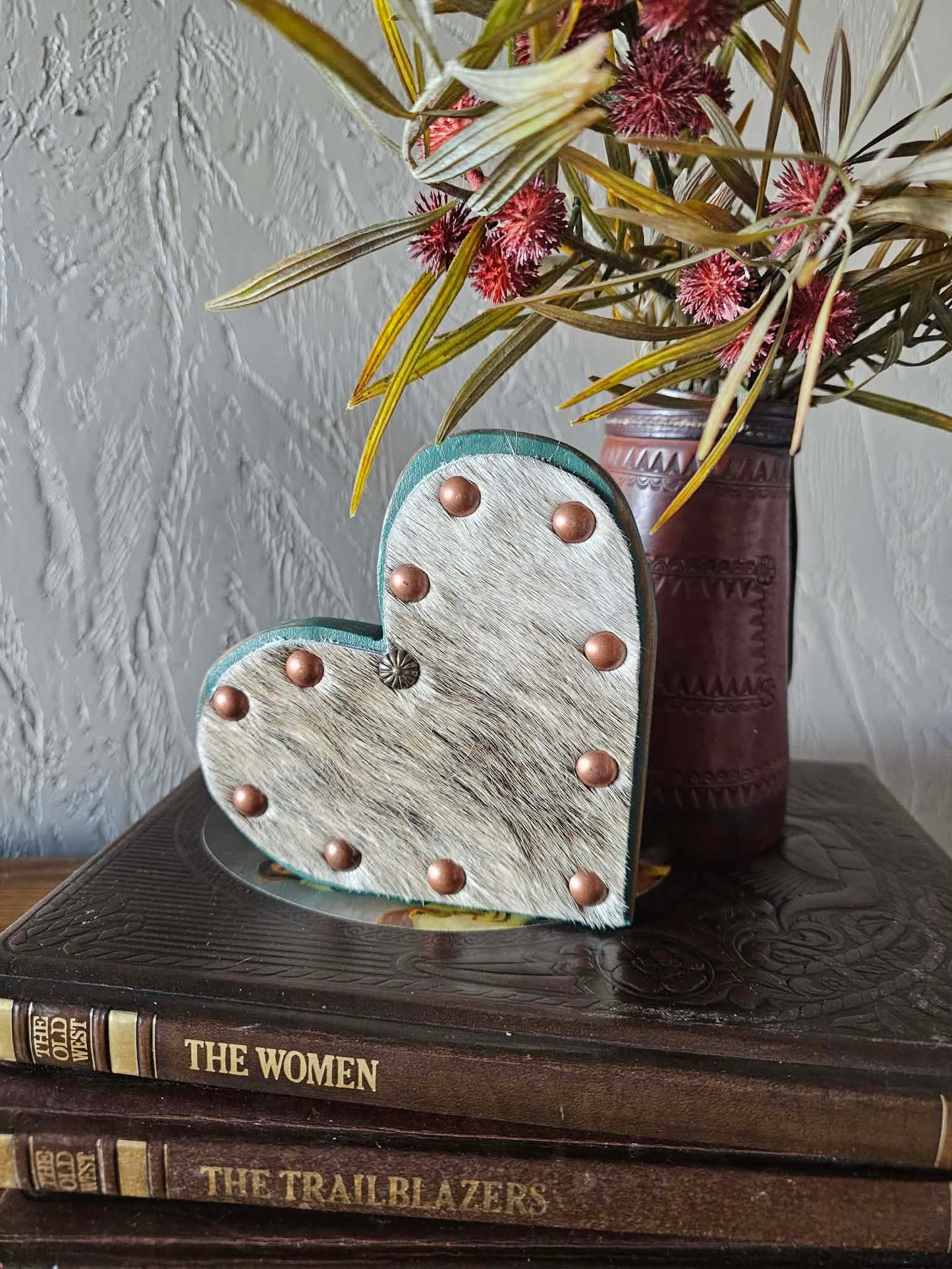 Heart-shaped decorative item with metallic accents on a stack of books, with a vase of flowers in the background.